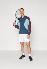 Chaqueta de cremallera azul y granate, pantalones cortos blancos, zapatillas deportivas negras. Sosteniendo una raqueta de tenis en una mano y una pelota de tenis en la otra.