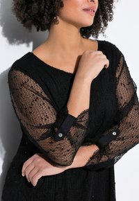 Top nero trasparente in maglia con maniche lunghe, caratterizzato da un motivo testurizzato e polsini solidi a contrasto con dettagli di bottoni.