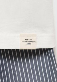 Hvid bomulds t-shirt med et mærke på kanten, der siger "MNG TEEN GENUINE GARMENTS 1984." Nedenunder er der mørkeblå stof med lodrette hvide striber.