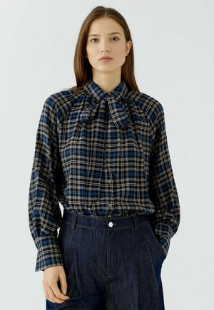 Frau, die eine blau-schwarz karierte Bluse mit Schleifenkragen und dunkle, hoch taillierte Jeans trägt, steht mit einer Hand in der Tasche.