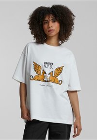 Hvit oversized t-skjorte med grafikk av to vingede skapninger i oransje og brunt, med teksten "BER N.Y.C." og andre trykte detaljer.
