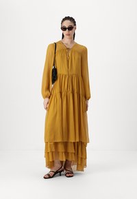 Vestido maxi amarelo mostarda com mangas longas, design em camadas, tecido leve e um laço subtil no decote. Sandálias pretas e uma bolsa complementam o visual.