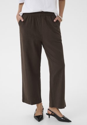 Kobieta ubrana w luźne luźne spodnie o szerokich nogawkach w ciemnobrązowym kolorze z elastycznym pasem, biały top oraz czarne czółenka na obcasie z odkrytą piętą, ręce w kieszeniach.