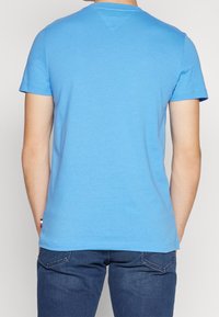 Muž v jednoduchém světle modrém tričku s krátkým rukávem a tmavě modrých džínách, pohled zezadu na jednobarevném pozadí.