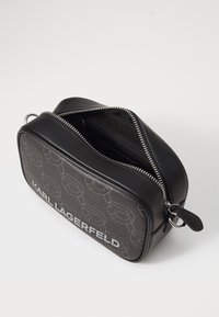 Črna usnjena torba z zadrgo, ki ima teksturirano stran z sivo oblogo in bold črkovanjem "KARL LAGERFELD" na sprednji strani.