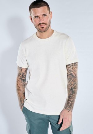Mann mit kurzen Haaren und Tätowierungen auf beiden Armen trägt ein schlichtes weißes Kurzarmshirt und grüne Hosen und steht vor einem hellen Hintergrund.