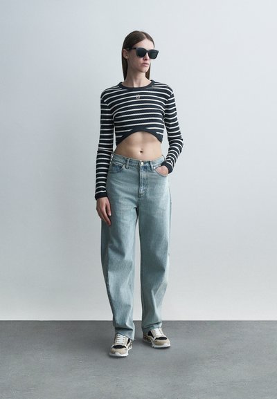 Γυναίκα που φοράει μαύρα γυαλιά ηλίου, μαύρο-άσπρο ριγέ κολλητό τοπ, τζιν ψηλόμεση ανοιχτού μπλε χρώματος και αθλητικά παπούτσια, στέκεται μπροστά σε απλό τοίχο.