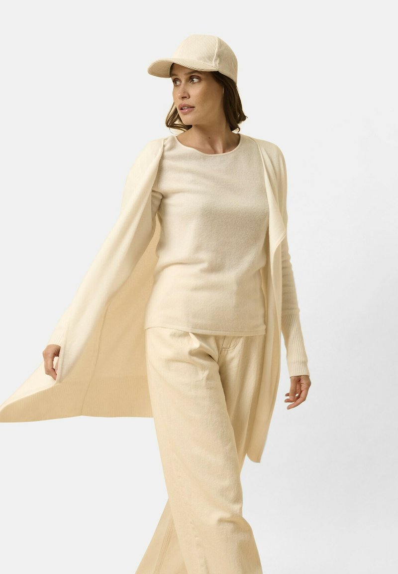 Suéter de punto beige, pantalones a juego y una gorra. El conjunto presenta un ajuste relajado con textura acanalada y mangas largas, promoviendo la comodidad.