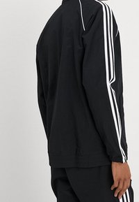 Person som bär en svart träningsoveralljacka med vita ränder längs ärmarna mot en enkel grå bakgrund.