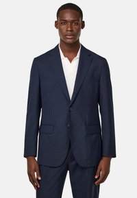 Blazer bleu marine avec une coupe ajustée, doté de deux poches avant, de revers à crans et de deux boutons. Porté sur un polo blanc.