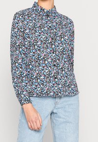 Blomstret bluse med sort baggrund, prydet med blå og pink mønstre, langærmet og med krave. Bæres med lyseblå jeans.