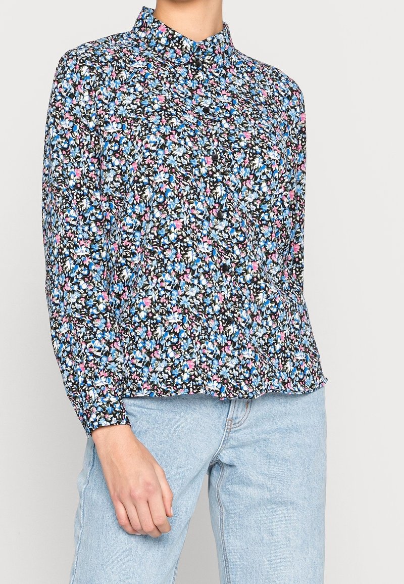 Blomstret bluse med sort baggrund, prydet med blå og pink mønstre, langærmet og med krave. Bæres med lyseblå jeans.