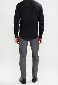 Man in een zwarte lange mouwen shirt, grijze broek, en zwarte nette schoenen, staande met zijn rug naar de camera tegen een effen witte achtergrond.