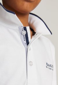 Nahaufnahme einer Person, die ein weißes Poloshirt mit navyfarbenem Besatz am Kragen und dem Logo "BAKER by Ted Baker" auf der Brust trägt.