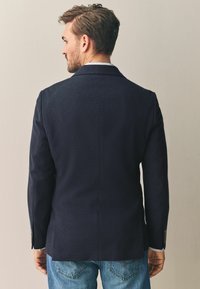 Tmavě modrý blazer se šitým střihem, strukturovanými rameny a úzkými klopami. Texturovaná tkanina s jemnými detaily a ladícími knoflíky.