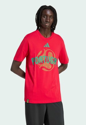 Persoon met dreadlocks, gekleed in een rood Portugal Adidas T-shirt en zwarte broek, staand met handen achter de rug tegen een effen achtergrond.