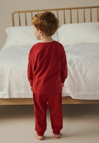 Cămașă roșie din bumbac cu mâneci lungi și pantaloni de pijama pe un copil, cu manșete și guler ribbate. Pat din lemn și lenjerie de pat albă în fundal.