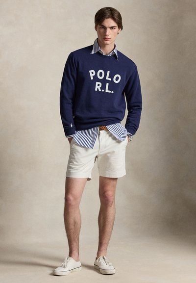 Jeune homme portant un sweat-shirt bleu marine « POLO R.L. » par-dessus une chemise rayée bleue et blanche, un short blanc et des baskets blanches, debout avec les mains dans les poches.