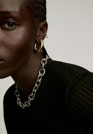 Pendiente de aro dorado y collar de cadena plateada lucidos sobre una blusa negra de canalé. Vista en primer plano del rostro, destacando los rasgos faciales y los accesorios.