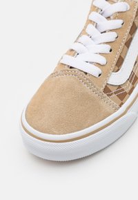 Zapatilla de ante beige con cordones blancos, detalles de cuadros en los costados, suela de goma y puntera texturizada.