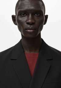 Blazer nero con risvolti decisi sopra un maglione rosso strutturato. Tessuto liscio con linee pulite, senza motivi visibili o decorazioni.