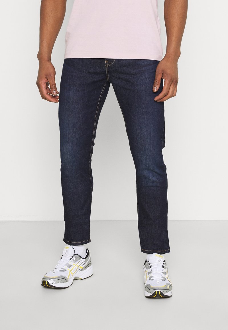 Homme portant un jean slim bleu foncé, des chaussettes blanches et des baskets blanches argentées avec des touches jaunes, debout devant un fond uni.