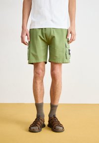Grüne Cargo-Shorts mit seitlichen Taschen, aus Baumwollstoff und mit lockerer Passform, kombiniert mit braunen Sandalen und grauen Socken. Strukturiert Oberfläche Details.