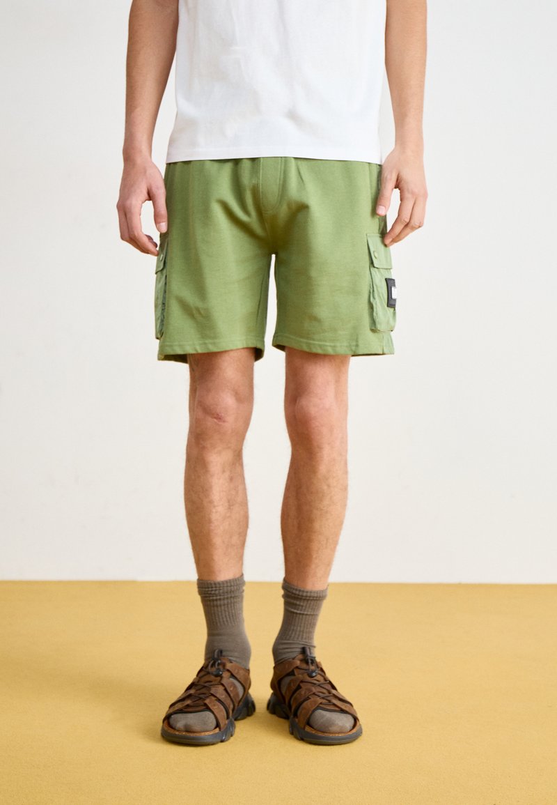 Grüne Cargo-Shorts mit seitlichen Taschen, aus Baumwollstoff und mit lockerer Passform, kombiniert mit braunen Sandalen und grauen Socken. Strukturiert Oberfläche Details.