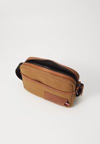 Brun lerret crossbody-veske med lærdetaljer, glidelåslukking, frontlomme og et lite farget merke på siden. Glatt tekstur.