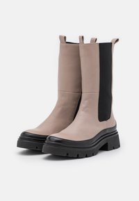 Beige leather ankle boots with black elastic side panels. Chunky black rubber soles with a shiny black toe cap. Two pulls at the top.