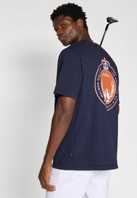 Tumši zili kokvilnas t-krekli ar lielu oranžu un baltu logotipu uz muguras, apaļš kakls un īsās piedurknes. Redzams golfa klubs.