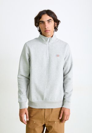 Light gray zip-up sweatshirt with a high collar, ribbed cuffs, and hem, featuring a small logo patch on the left chest. Smooth texture.