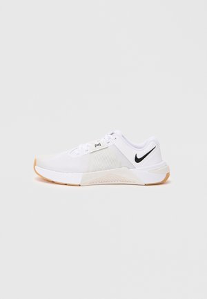 Hvid Nike sportssko med sort swoosh-logo, mesh-overdel og gummisål, vist fra ydersiden på en ensfarvet lys baggrund.