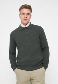 Polo en tricot vert foncé avec manches longues, patte de boutonnage à cinq boutons et poignets côtelés. Porté sur une chemise à col rose pâle.