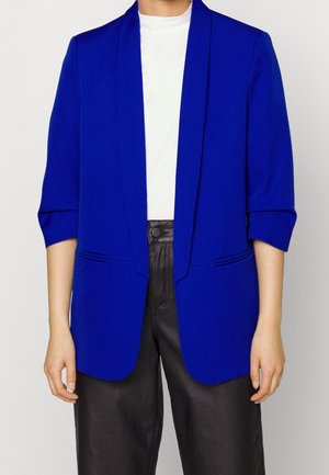 Persona con una chaqueta azul real sobre una camisa blanca y pantalones de cuero negros de talle alto, de pie con los brazos relajados a los lados.