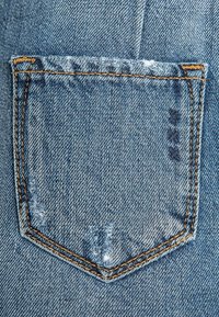 Bolsillo trasero de mezclilla con forma triangular, bordes deshilachados, costura naranja y un ligero desgaste, presentado sobre un fondo de tela azul texturizada.