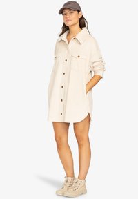 Robe chemise en velours côtelé beige à manches longues, deux poches poitrine, boutons marron, fentes sur les côtés, portée avec des bottes beiges à lacets et une casquette.