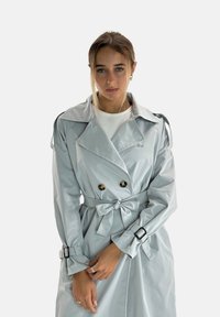 Trench coat grigio chiaro con ampi revers, design doppiopetto, due grandi bottoni, cintura regolabile e dettagli con fibbia sui polsini delle maniche.