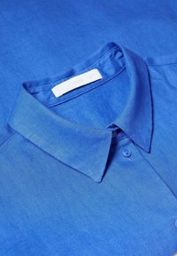 Chemise bleue à col avec des boutons et une étiquette blanche visible à l’intérieur du col.