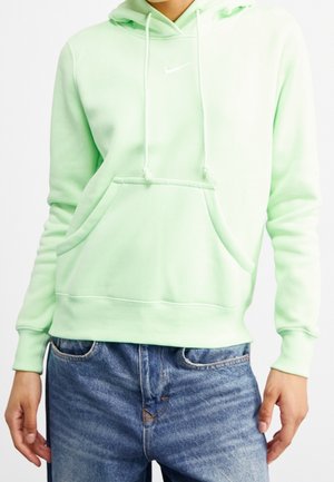 Personne portant un sweat à capuche Nike vert clair avec poche avant et cordons, associé à un jean bleu sur un fond uni.