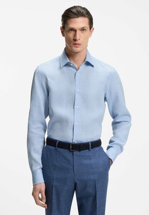 Mann, der ein hellblaues langärmeliges Hemd trägt, in blaue, maßgeschneiderte Hosen mit einem schwarzen Gürtel gesteckt, posiert mit einer Hand in der Tasche.