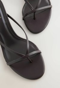 Gros plan sur des sandales en cuir noir minimalistes avec de fines brides entrecroisées et un design de bout carré sur fond blanc.