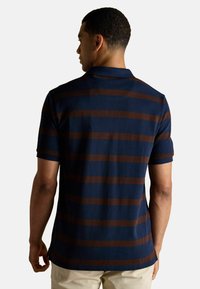 Jūras spēku polo krekls ar horizontālām brūnām svītrām, īsām piedurknēm, apkakli un sānu izgriezumiem apakšmalā. Mīksta adīta auduma tekstūra.