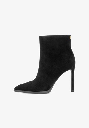 Czarne zamszowe botki za kostkę z ostrym czubkiem i wysokim obcasem w stylu stiletto. Charakteryzują się minimalistycznym designem i subtelnym akcentem w postaci złotego logo.