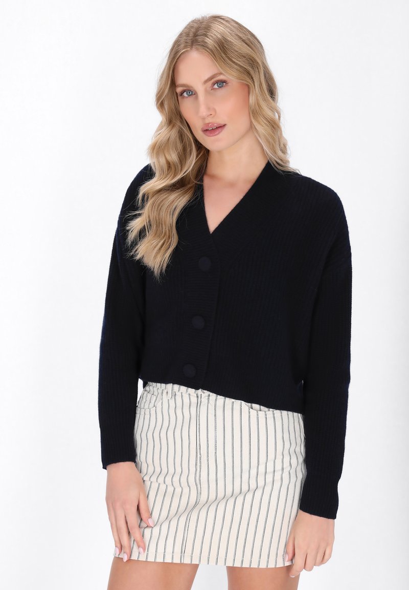 Cardigan cropped blu navy in maglia a coste con scollatura a V, caratterizzato da tre grandi bottoni e maniche lunghe, abbinato a una gonna a righe.