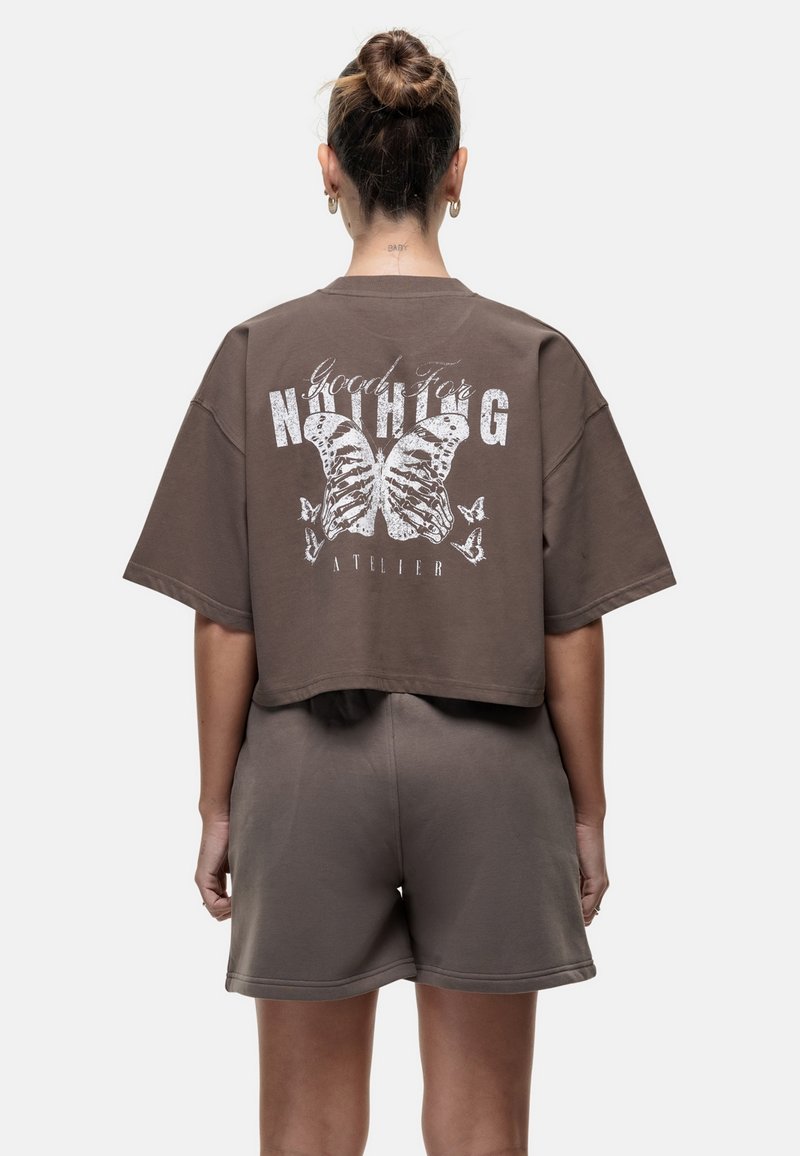 Bruine cropped T-shirt met een witte vlinderafbeelding en tekst op de achterkant. In combinatie met bijpassende bruine shorts. Zacht materiaal, losse pasvorm.