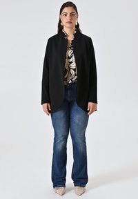 Schwarzer langer Mantel mit minimalistischer Gestaltung, getragen über einer gemusterten cremefarbenen Bluse. Kombiniert mit blauen Jeans und hellbeigen spitzen Schuhen.