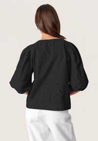 Top nero con maniche a palloncino e scollatura ampia, con dettagli intagliati ricamati lungo l'orlo. Tessuto morbido, abbinato a pantaloni bianchi.