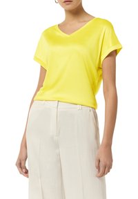 Żółta koszulka z dekoltem w serek, z gładkiego materiału, z krótkimi, podwiniętymi rękawami, zestawiona z jasnymi, plisowanymi spodniami.