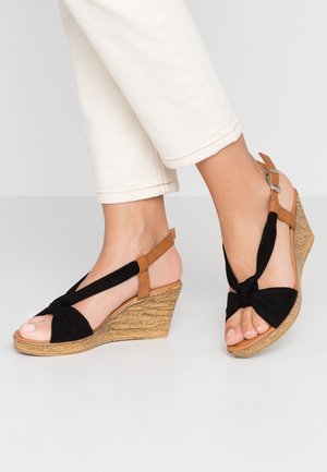 Sorte kilehæle med knyttet sort stofoverdel, brune ankelremme og struktureret beige kilehæl, præsenteret med hvide cropped bukser.